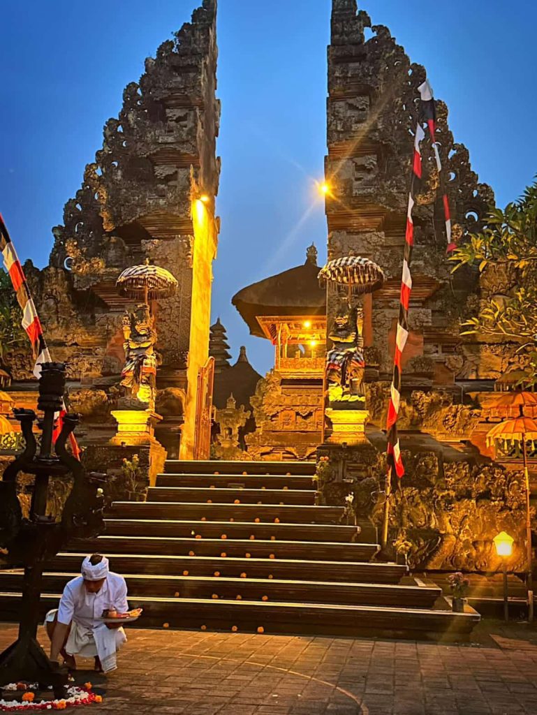 Balinese man offers prayers at a temple gate adorned with flags and lanterns.