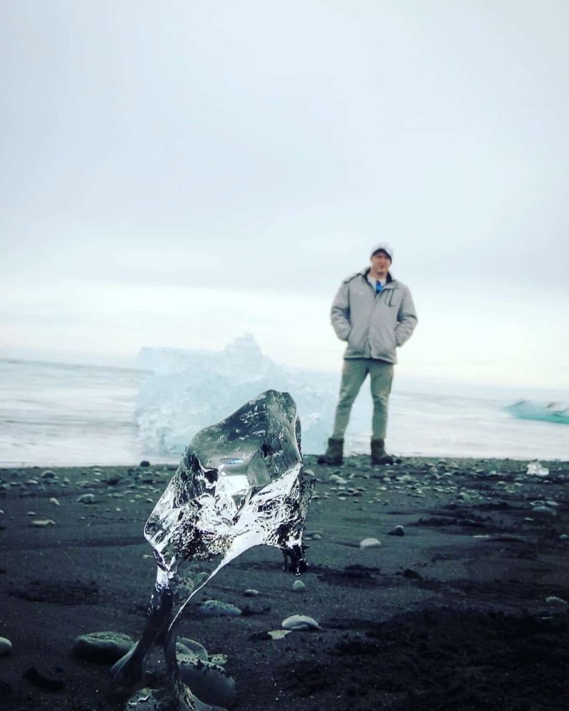 Man stands on black sand beach in Iceland with ice formation in foreground. Reykjavik travel photo.