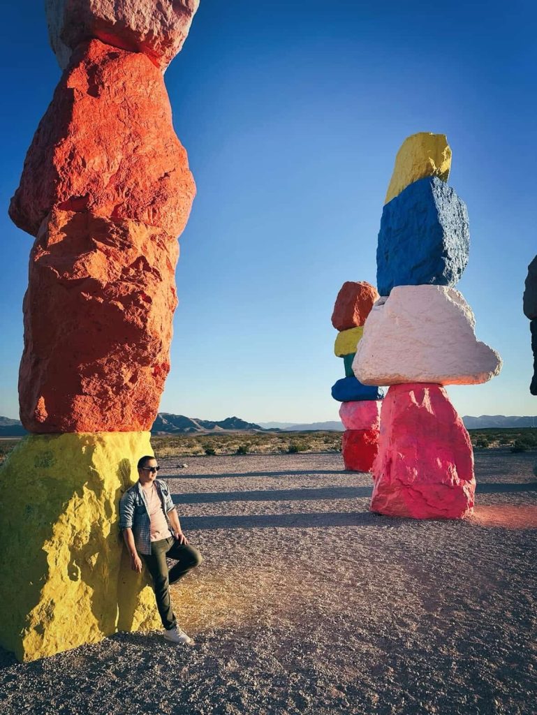Man at Seven Magic Mountains art installation near Las Vegas, Nevada. Colorful rock sculptures against a clear blue sky.