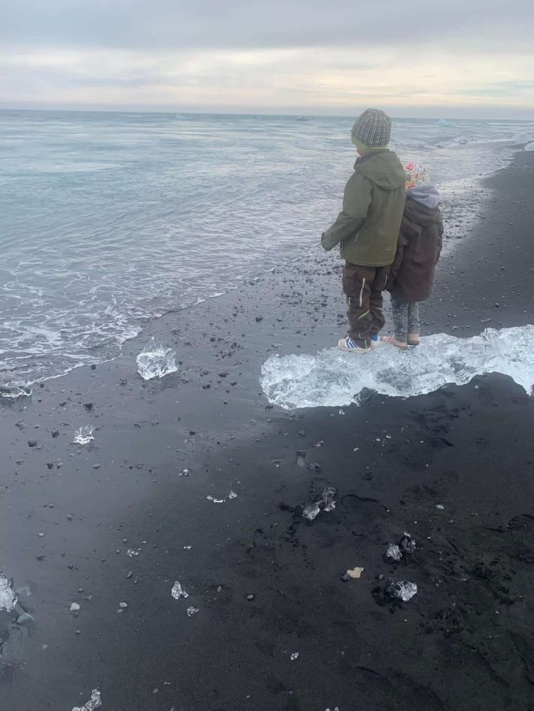 Two children stand on an iceberg on a black sand beach in Reykjavik, Iceland, gazing at the ocean.