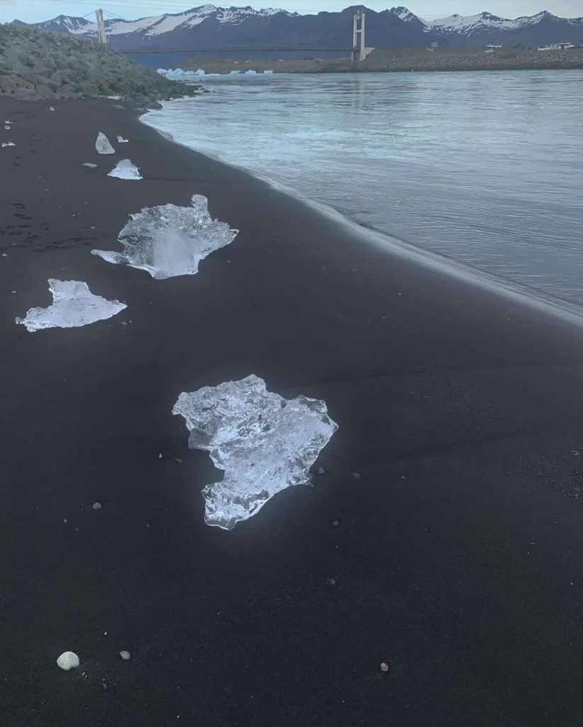 Ice chunks on black sand beach near Reykjavik, Iceland, with bridge and mountains in background.