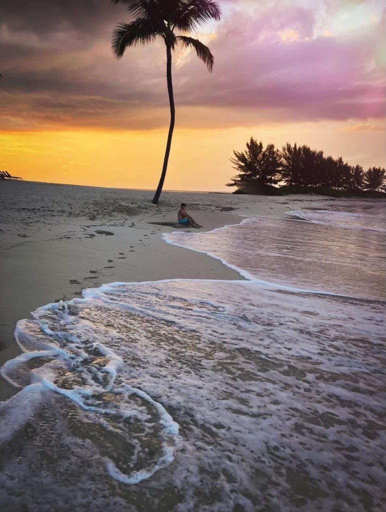 Person sitting on a beach in Nassau at sunset with palm tree and ocean waves.
