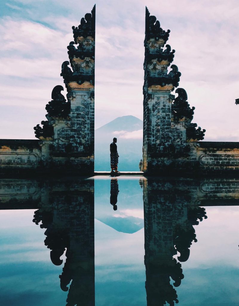 Silhouette of a person standing at the Lempuyang Temple gates in Bali, with a volcano reflection.
