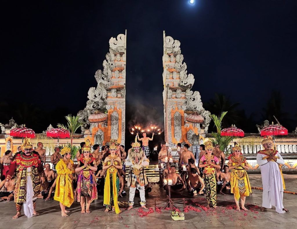 Balinese dancers in traditional costumes perform at night in front of a temple gate.