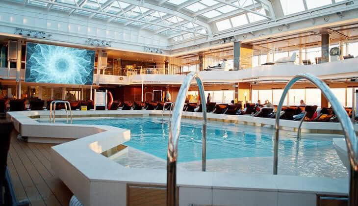 Cruise ship pool with skylight and lounge chairs.