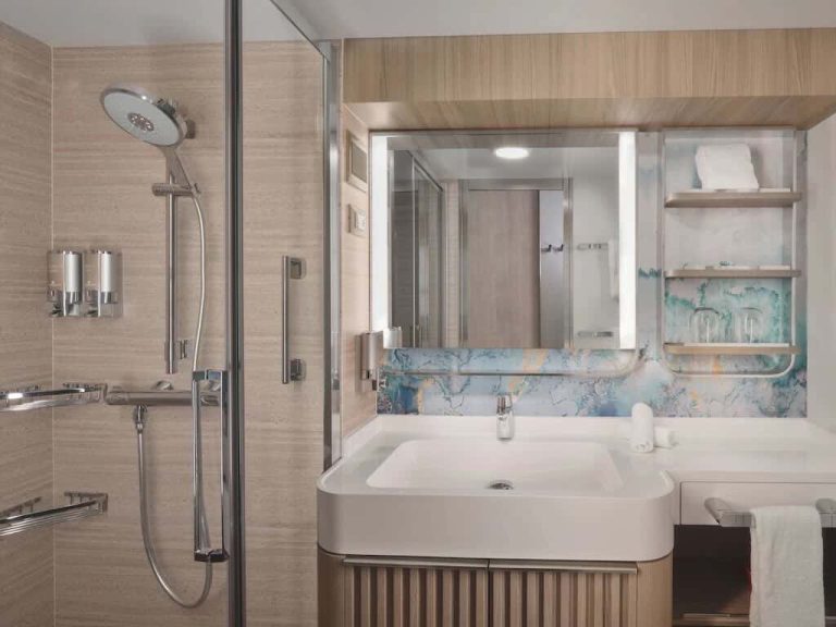 Cruise ship bathroom with shower, sink, and storage. Modern design with light wood and marble-look walls.