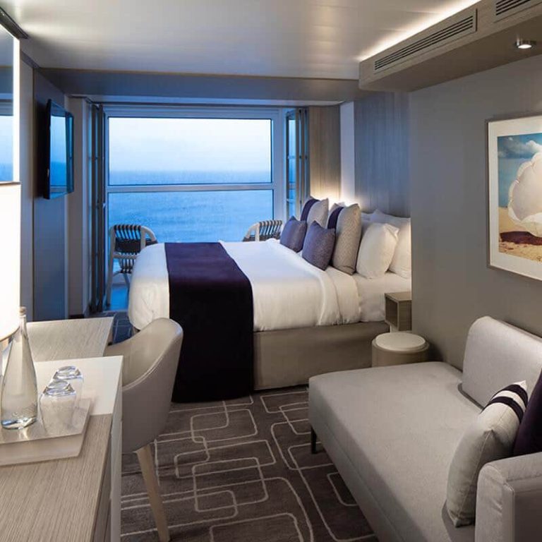 Cruise stateroom with balcony overlooking the ocean, featuring a bed, sofa, and desk.