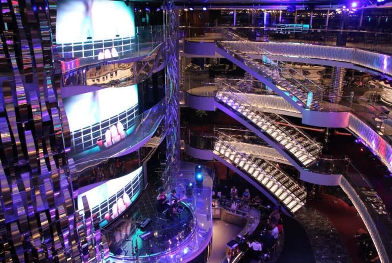 Cruise ship atrium with escalators, bar, and LED screens.