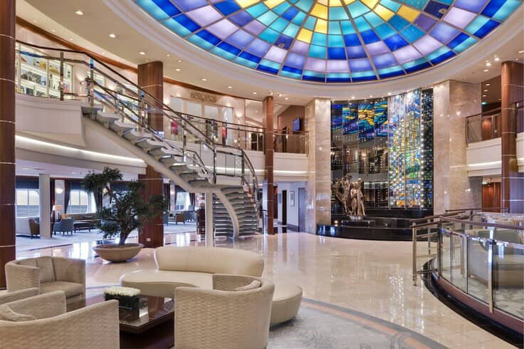 Cruise ship atrium with grand staircase, dome ceiling, and elegant seating area.