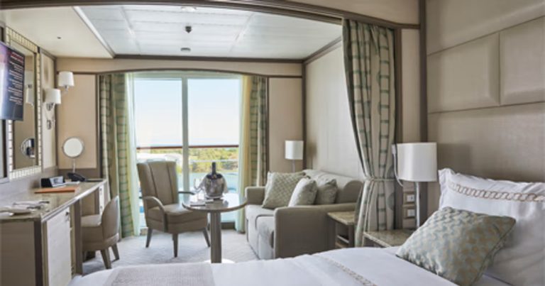 Cruise ship cabin with balcony, sofa, desk, and bed. Neutral color scheme.