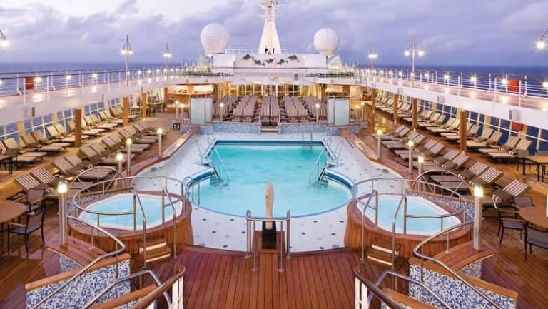 Cruise ship deck with pool, hot tubs, and lounge chairs under soft lighting.