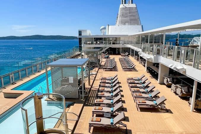 Cruise ship deck with lounge chairs and swimming pool under blue sky.