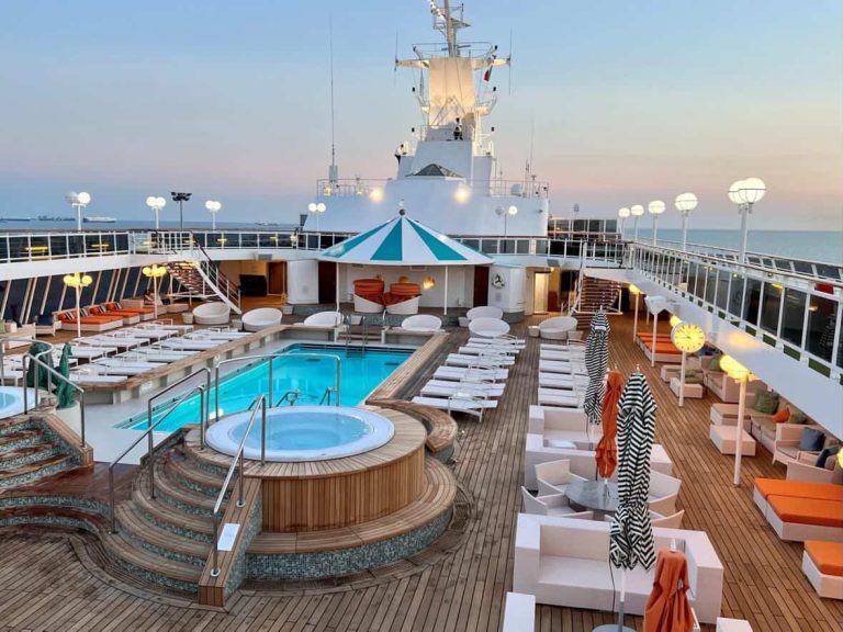 Cruise ship deck with pool, hot tub, lounge chairs at sunset.