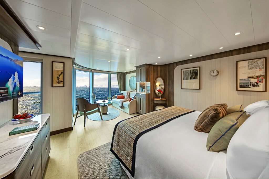Luxury cruise suite with ocean view, bed, seating area, and TV displaying icy landscape.
