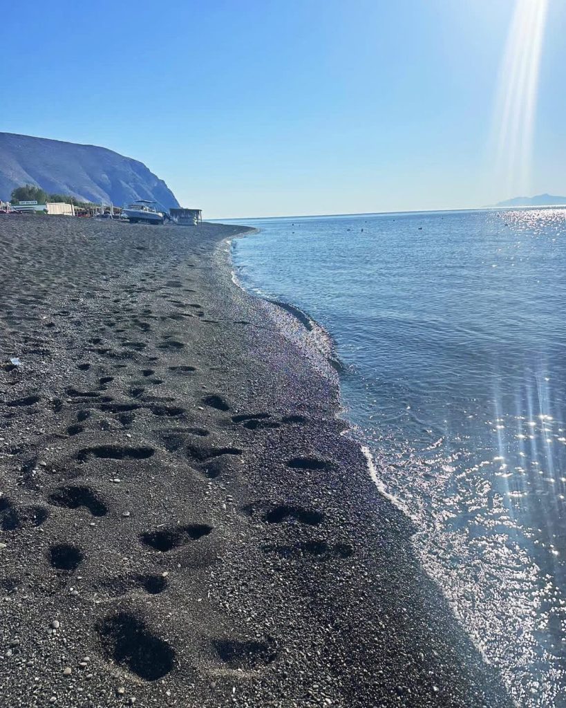 Black sand beach with footprints and calm blue ocean under a bright sunny sky.