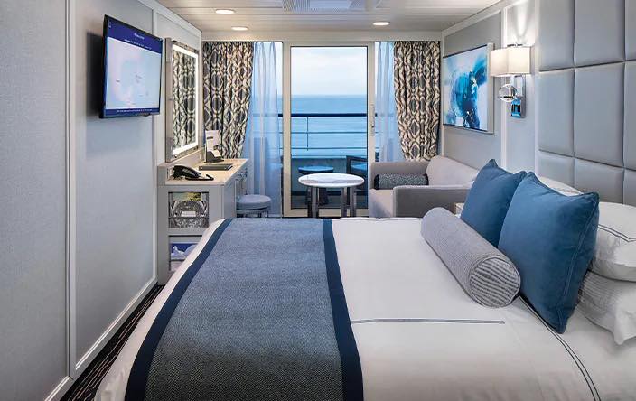 Modern cruise ship cabin with a king bed, desk, and large window overlooking the ocean.