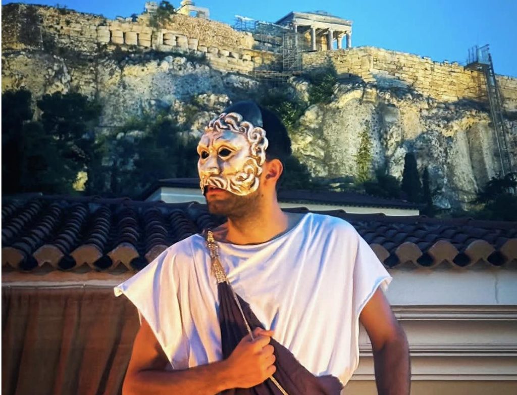Man in ancient Greek costume and mask stands before the Acropolis