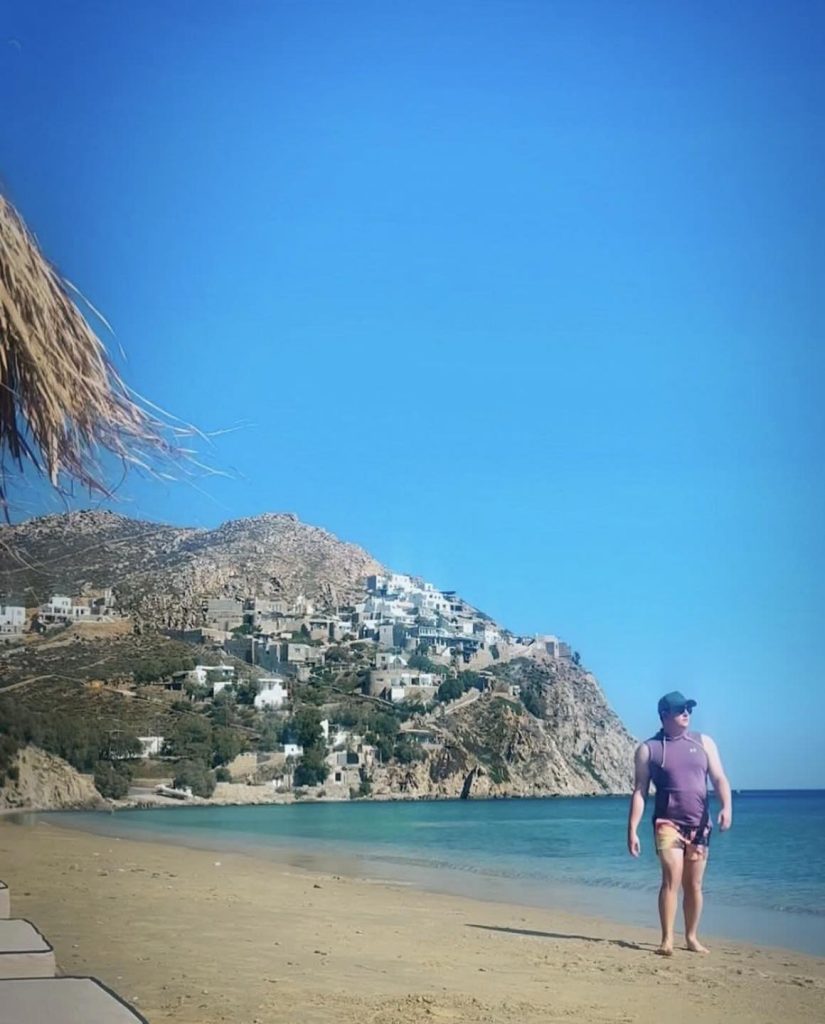 Man walks on a Greek island beach with a village on a hillside above.