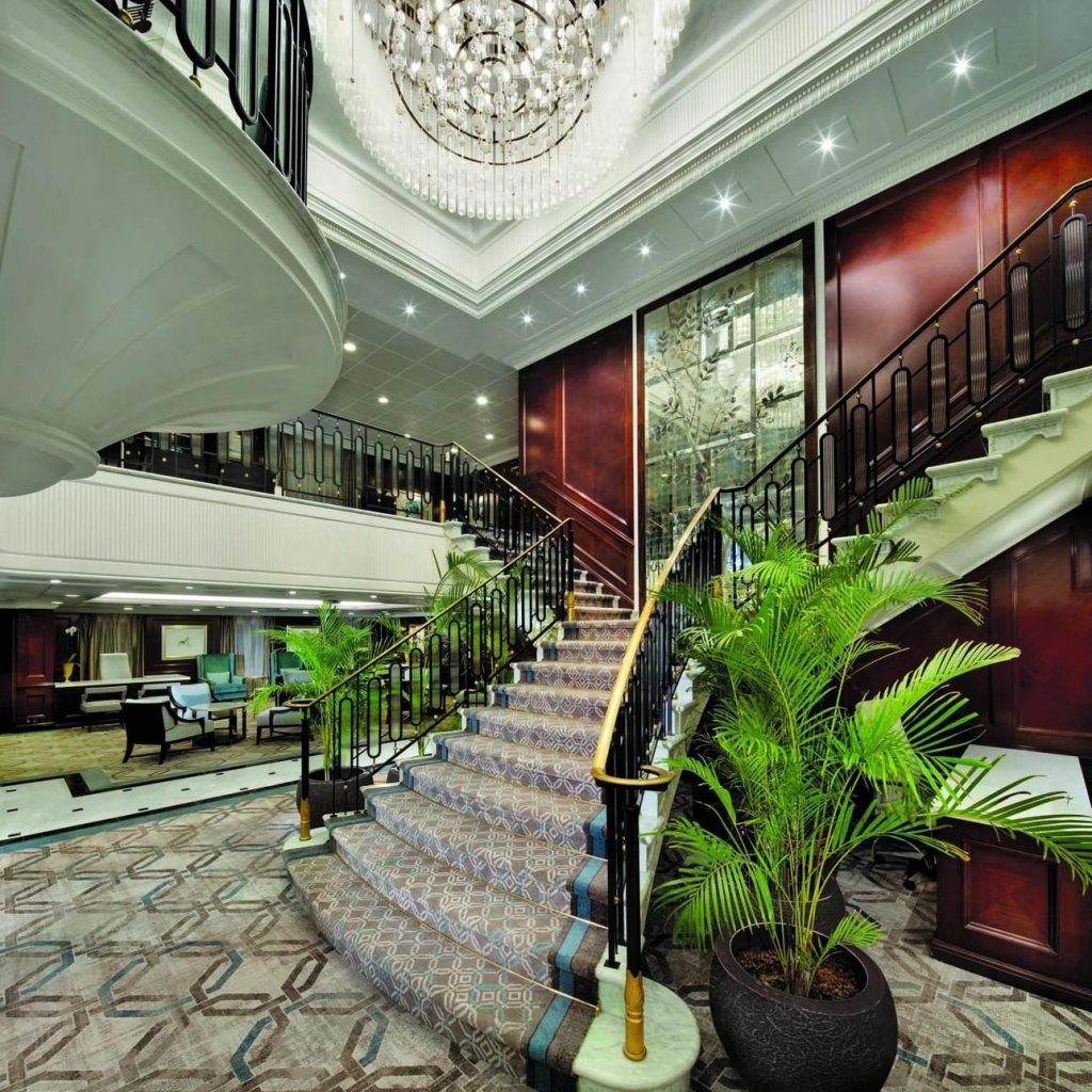 Luxurious hotel lobby with grand staircase, chandelier, and potted plants.