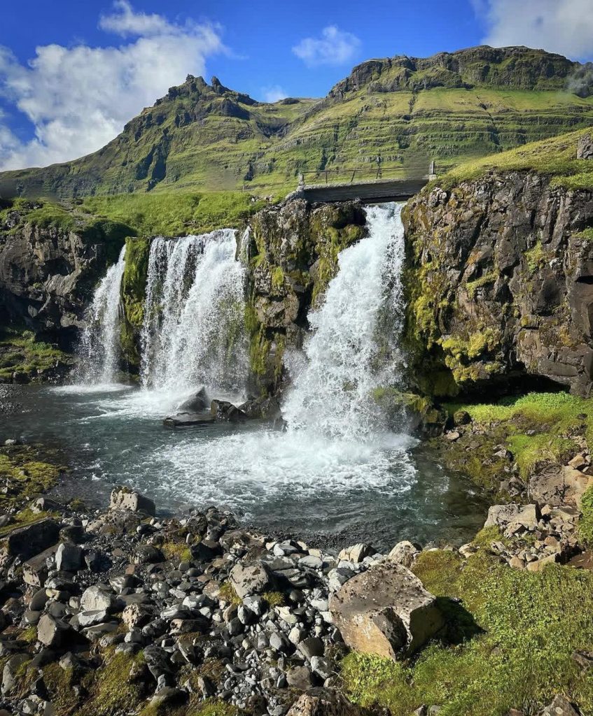 Cascading waterfalls flow over moss-covered rocks into a clear pool, framed by green mountains.