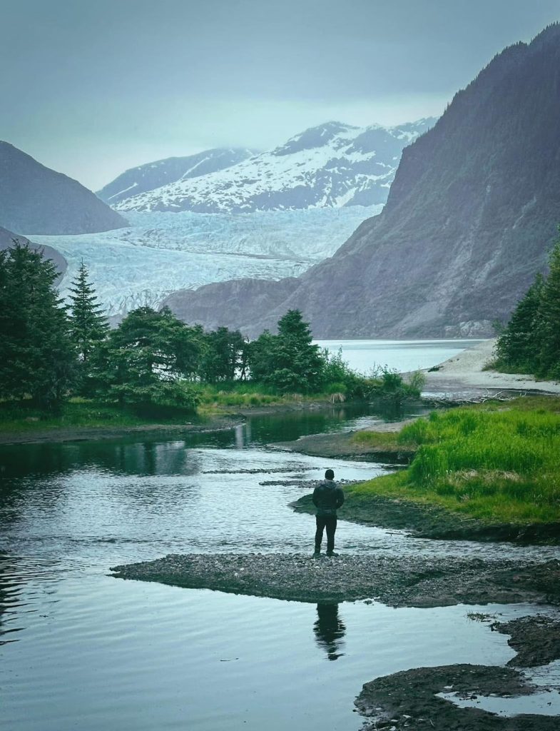 Man stands at the edge of a river with a glacier and mountains in the background.