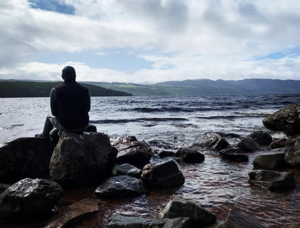 Man sits on rocks overlooking a vast, choppy lake with mountains in the distance.