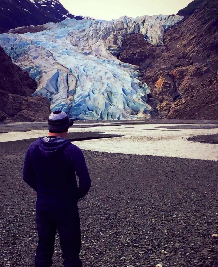 Man in purple hoodie faces a vast blue glacier in a rocky valley.